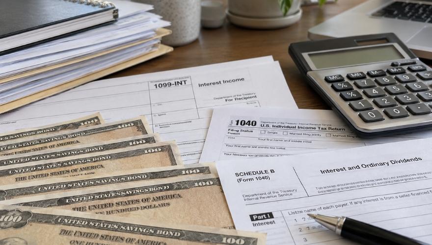 U.S. savings bonds displayed alongside IRS Forms 1099-INT, 1040, and Schedule B, illustrating how to report taxable savings bond interest on a federal tax return.